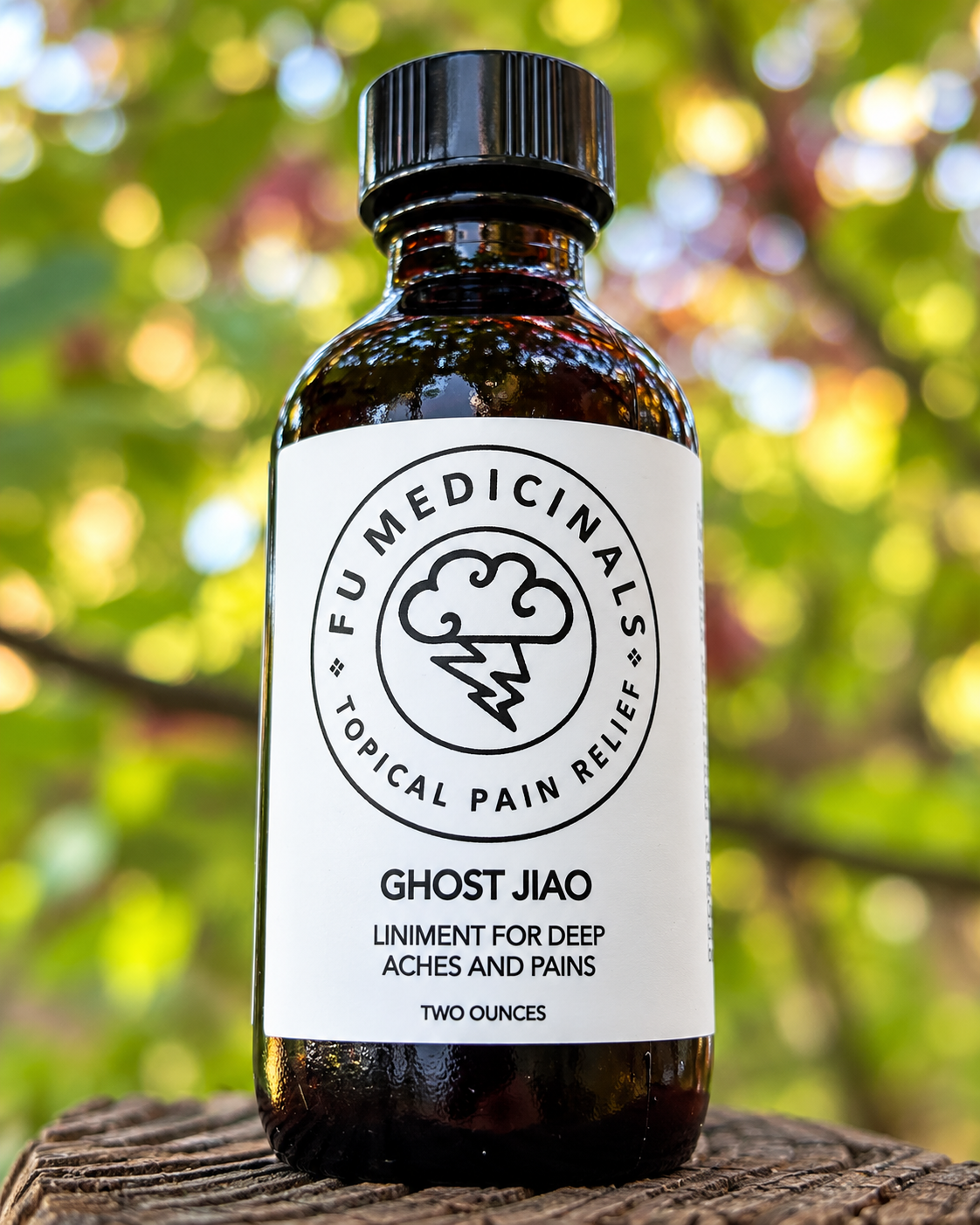 A bottle of FU Medicinals Ghost Jiao topical pain relief liniment, shown on a rustic wooden surface with a blurred green and yellow outdoor background.